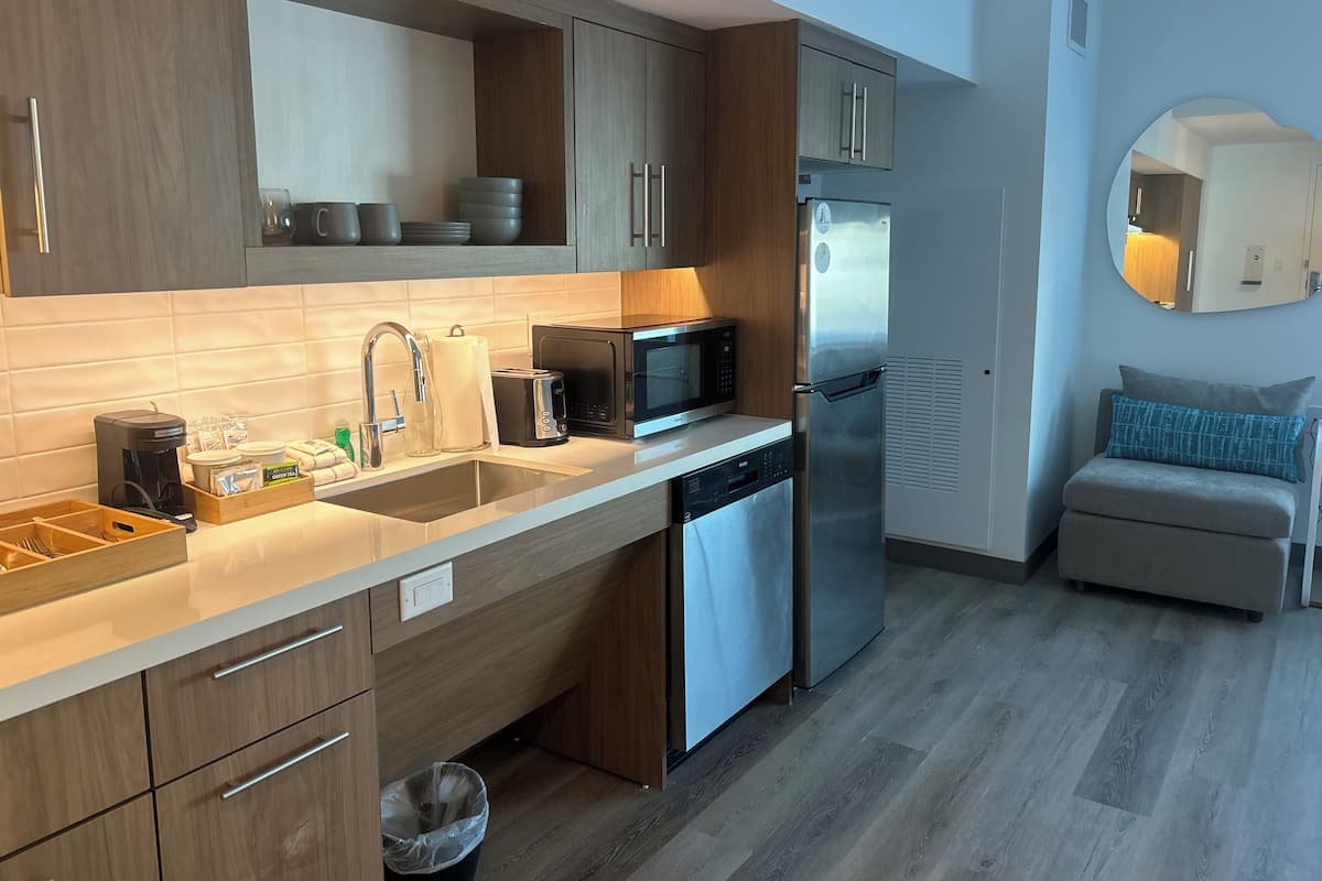 A modern kitchen with a stainless steel refrigerator, a microwave, and a dishwasher. There is a wooden cabinet above the sink and a small seating area with a cushion.