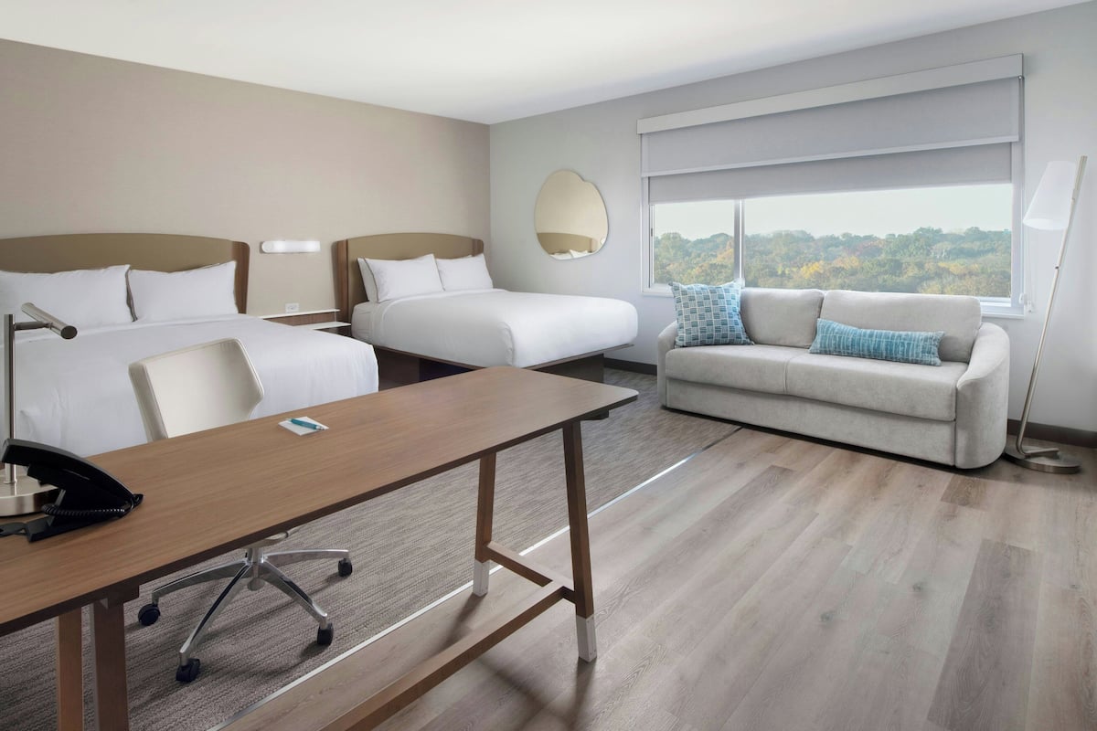 A hotel room with a wooden desk, two beds, a sofa, and a large window with a view.