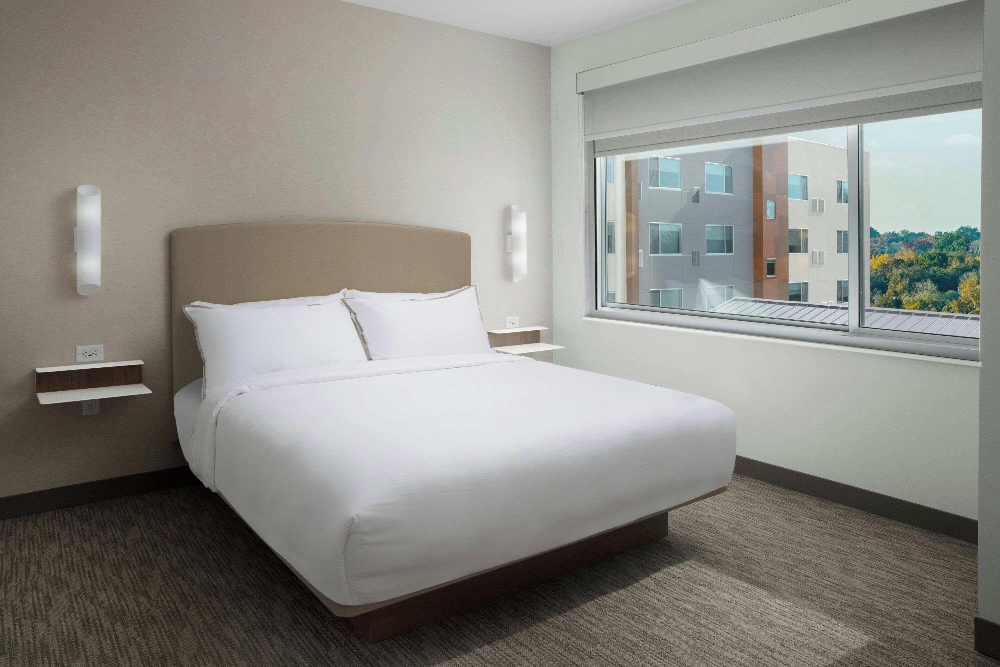 A hotel room with a large bed, a window with a city view, a bedside table, and a wall-mounted light.