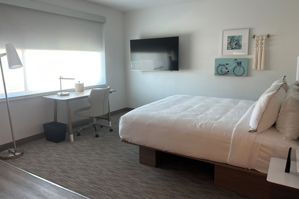 A modern hotel room with a large bed, a desk with a chair, a flat-screen TV, and artwork on the walls.