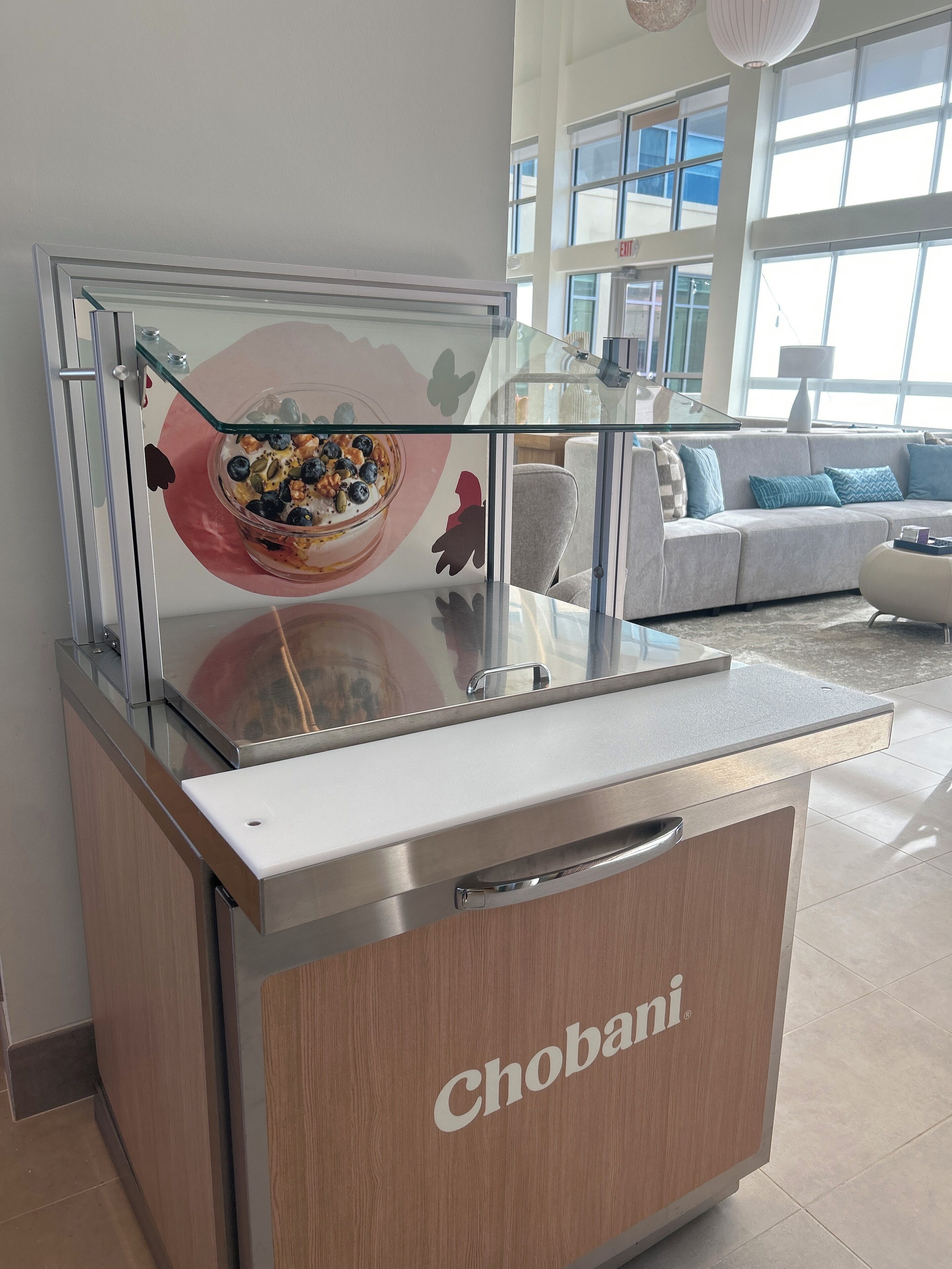 A Chobani yogurt display in a modern, well-lit room with a large window, a sofa, and a glass partition.
