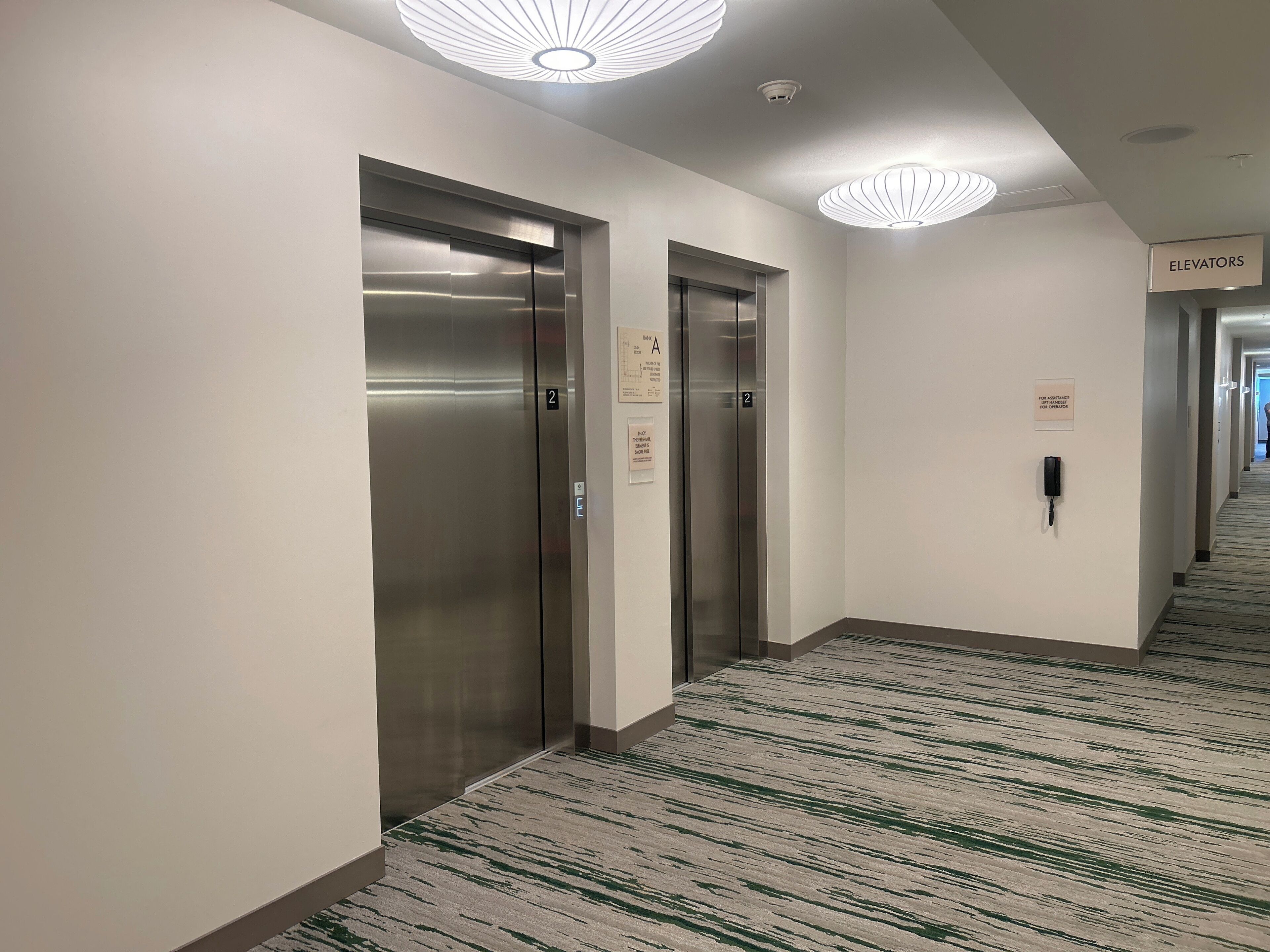 Un pasillo moderno de hotel con dos ascensores, piso alfombrado y un cartel que indica la ubicación de los ascensores.