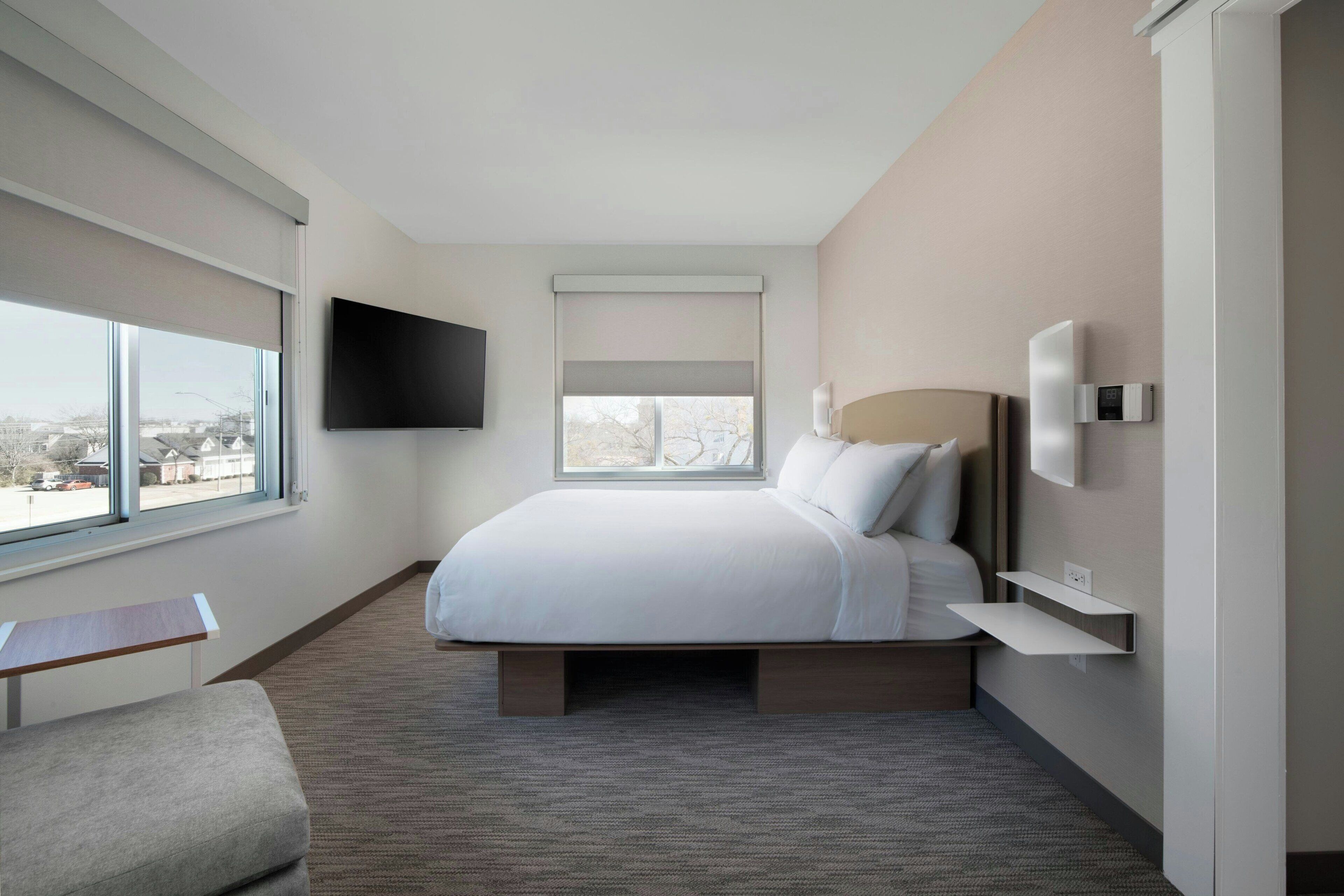 Une chambre d’hôtel moderne dotée d’un grand lit, d’un téléviseur à écran plat fixé au mur, d’une table de chevet et d’une fenêtre offrant une vue sur la ville.