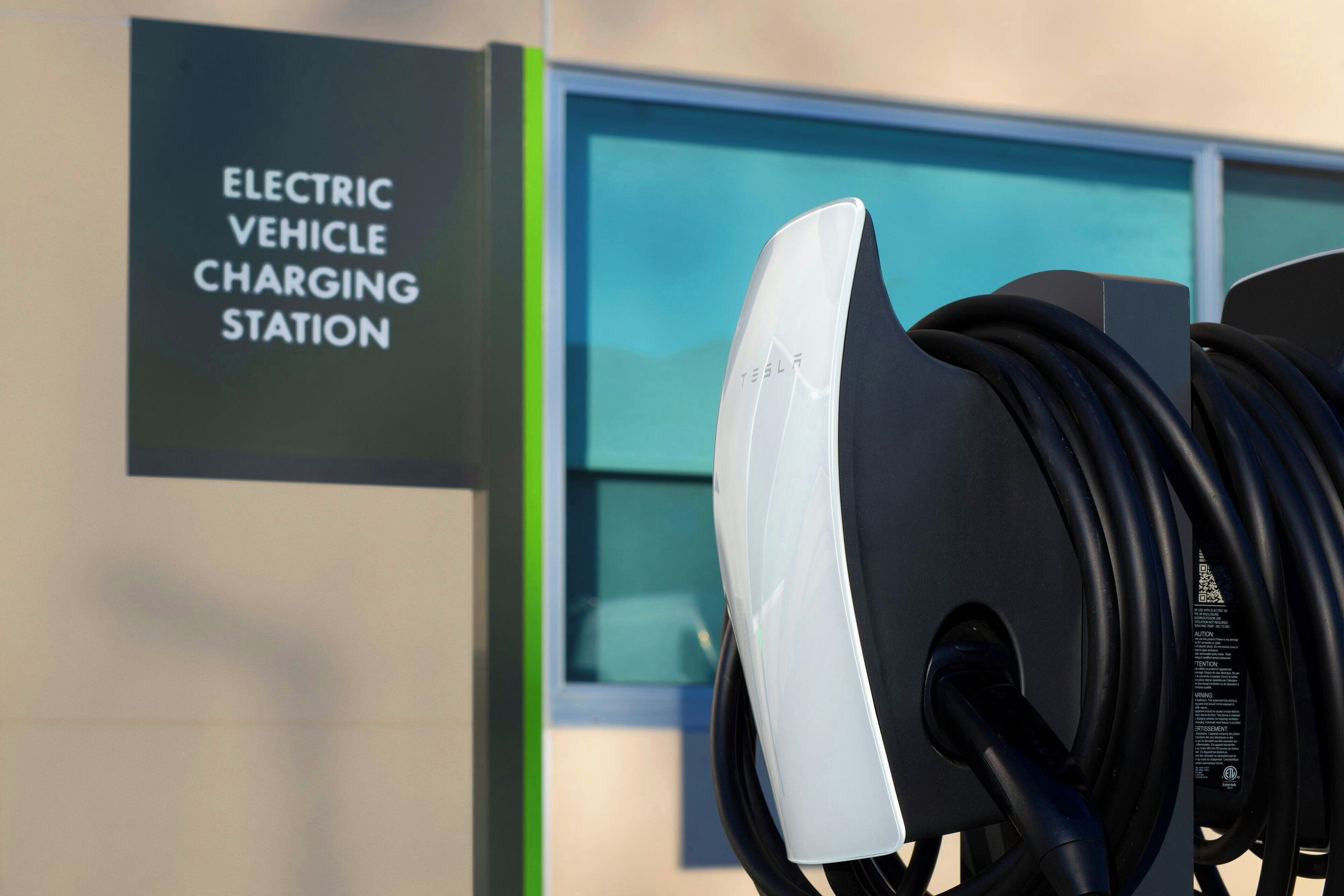 An electric vehicle charging station with a Tesla logo, a charging cable, and a sign indicating the station's purpose.