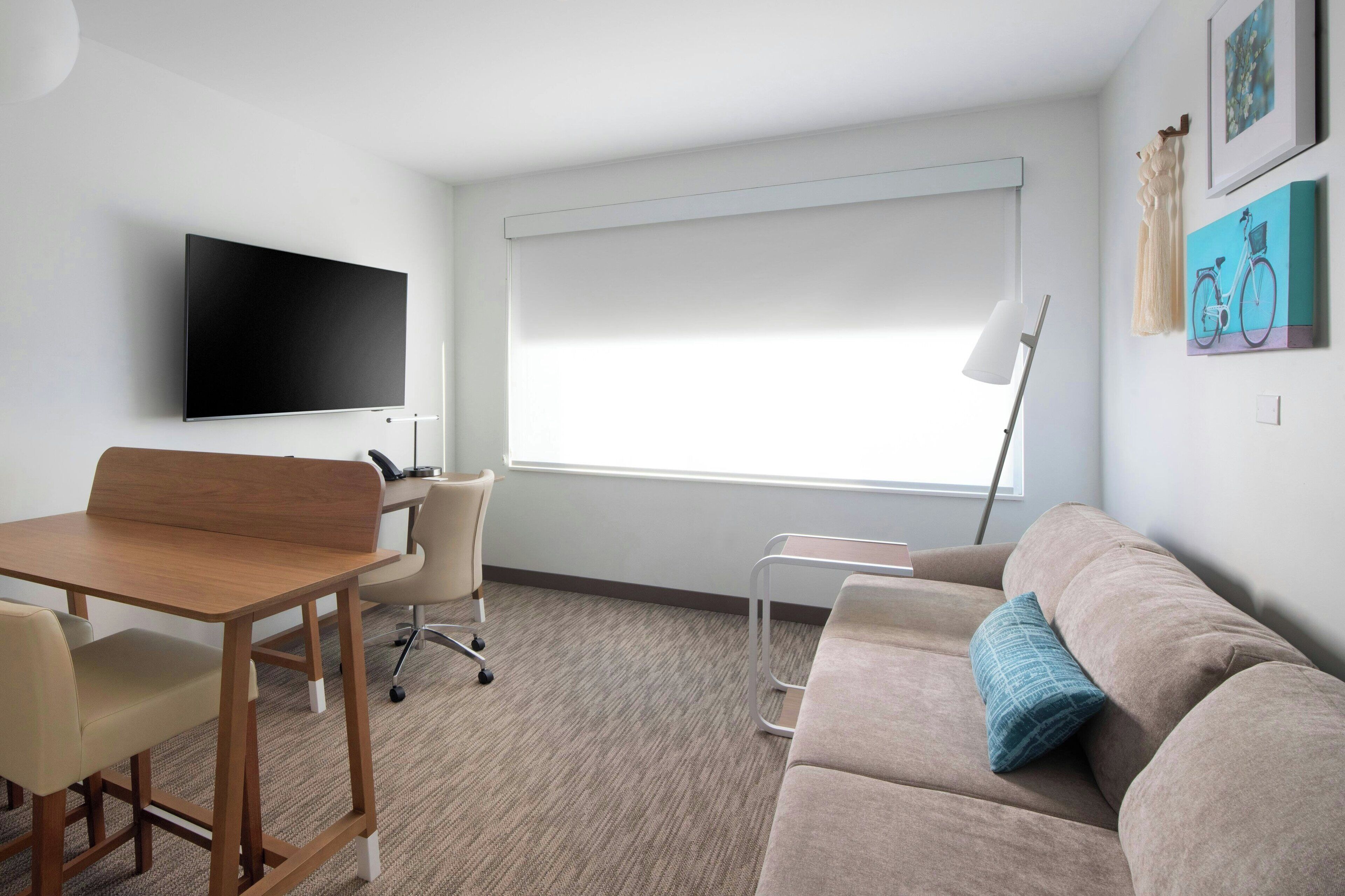 A modern room with a flat-screen TV, a desk, a chair, a sofa, and a window with blinds.