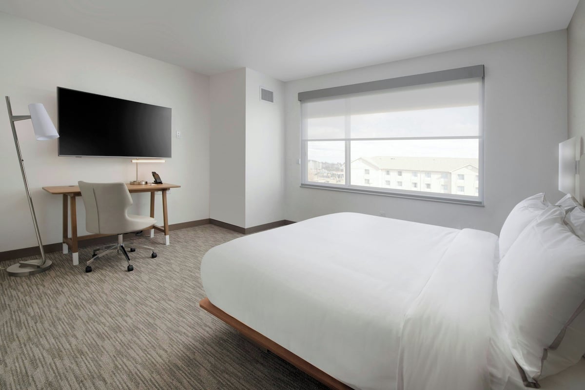 A hotel room with a bed, a desk with a chair, a TV mounted on the wall, and a window with a view of buildings.
