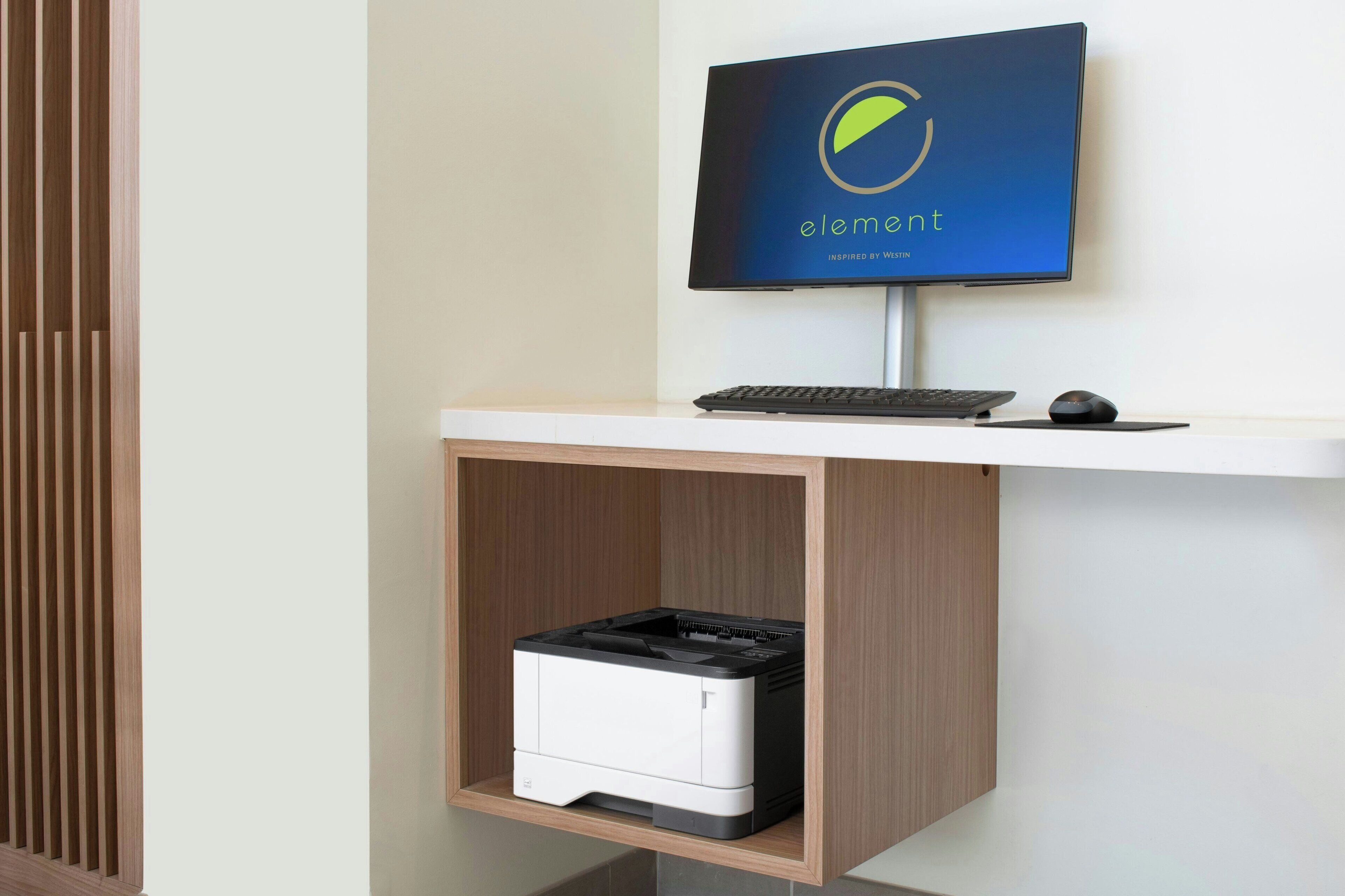 A modern office setup with a flat-screen monitor, keyboard, and mouse on a white desk, and a printer in a wooden cabinet below.