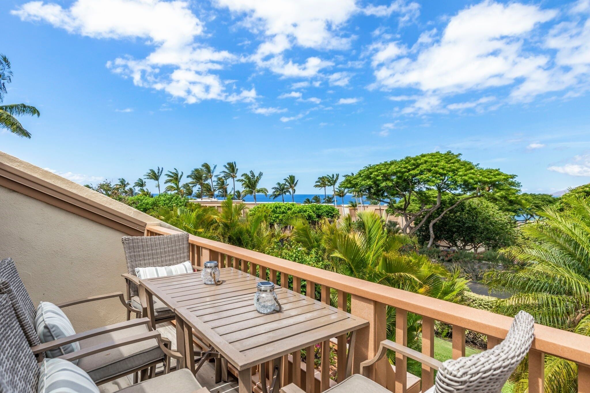 Una terraza de madera con mesa y sillas, con vistas a un paisaje tropical con palmeras y el mar.