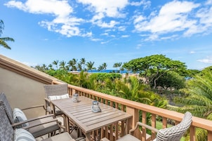 A wooden deck with a table and chairs, overlooking a tropical landscape with palm trees and a view of the ocean.