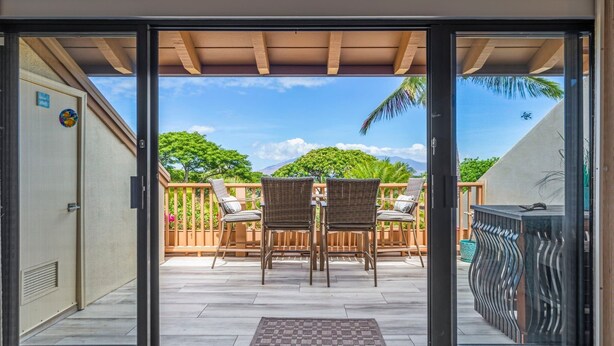 A sliding glass door leading to an outdoor patio with a wooden deck, wicker chairs, and a view of a mountain. - Maui Kamaole, #i215 2 Bedroom Condo by RedAwning (Kihei)