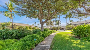 A pathway flanked by lush greenery and palm trees, leading to a building with a tiled roof. - Maui Kamaole, #i215 2 Bedroom Condo by RedAwning (Kihei)