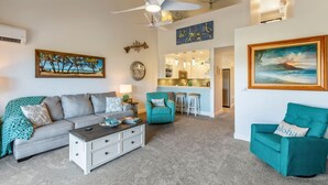 A living room with a sofa, a teal armchair, a coffee table, and a dining area with stools. - Maui Kamaole, #i215 2 Bedroom Condo by RedAwning (Kihei)