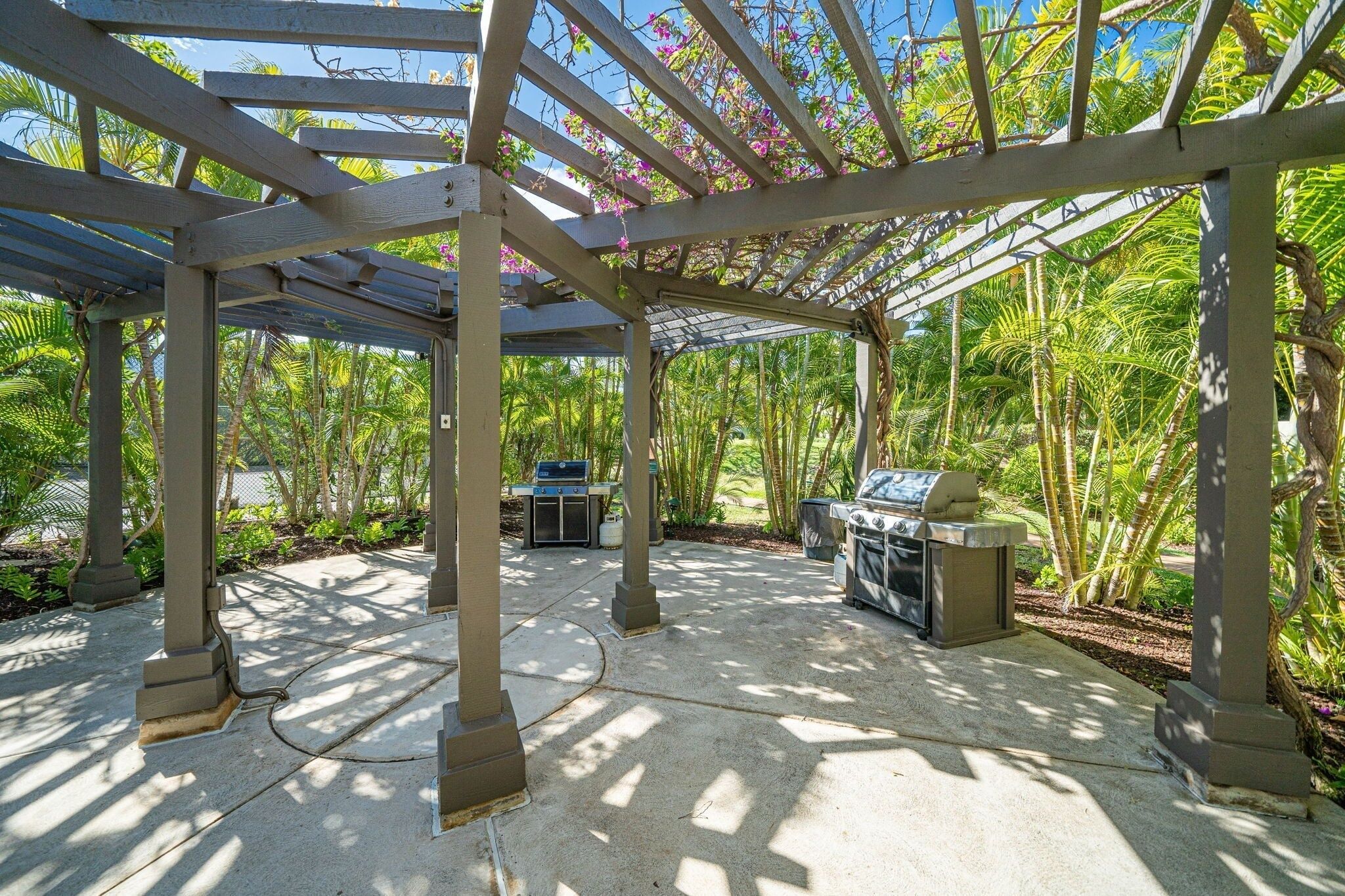 Un patio al aire libre con una pérgola, una parrilla de barbacoa y un área de estar rodeada de abundante vegetación.