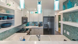 A modern kitchen with a central island, stainless steel appliances, and teal glass pendant lights. - Maui Kamaole, #i215 2 Bedroom Condo by RedAwning (Kihei)
