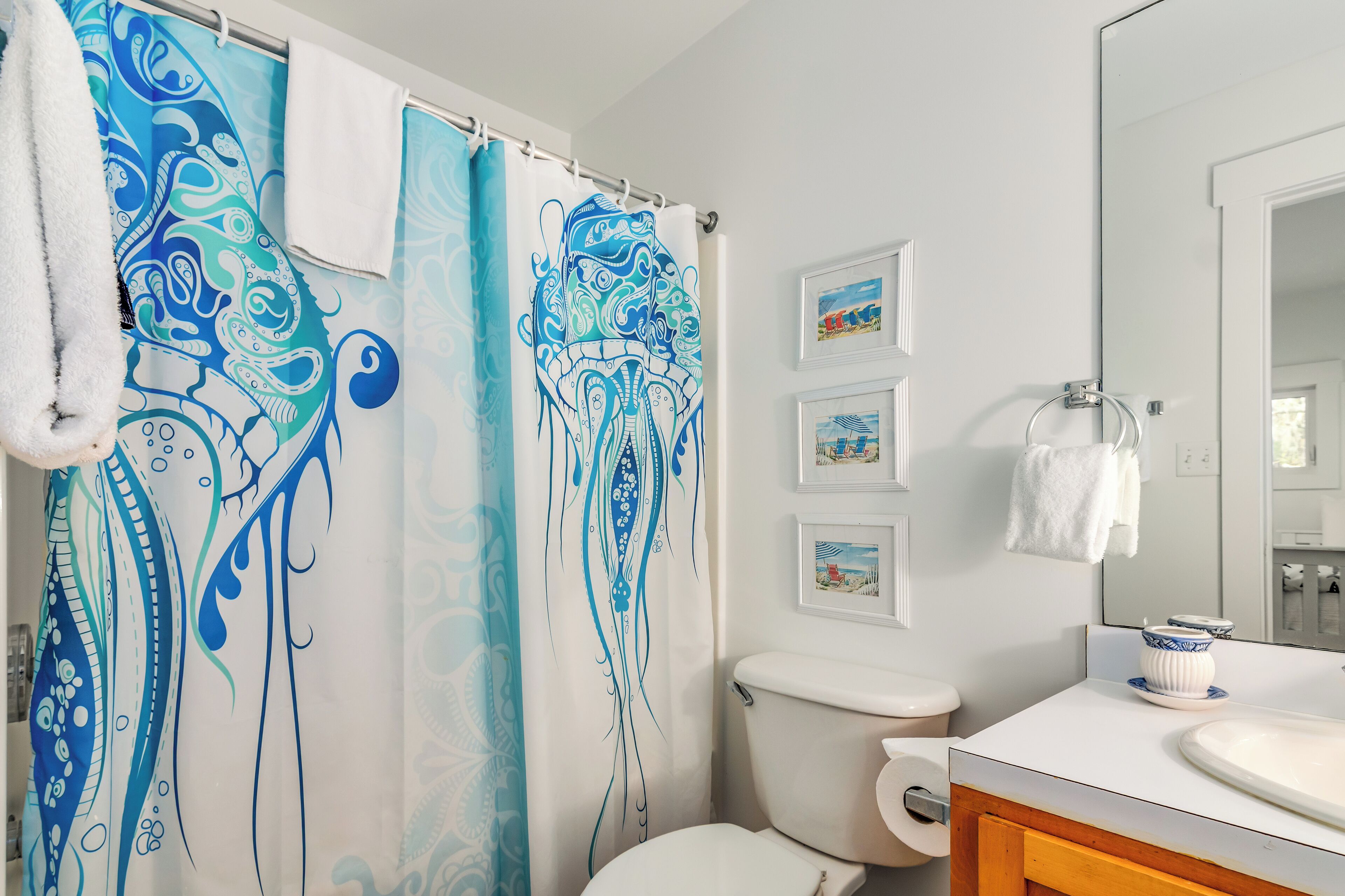 Une salle de bain avec un rideau de douche orné d’un motif abstrait bleu et blanc, des toilettes, un lavabo avec un plan de toilette blanc et un miroir au-dessus du lavabo.