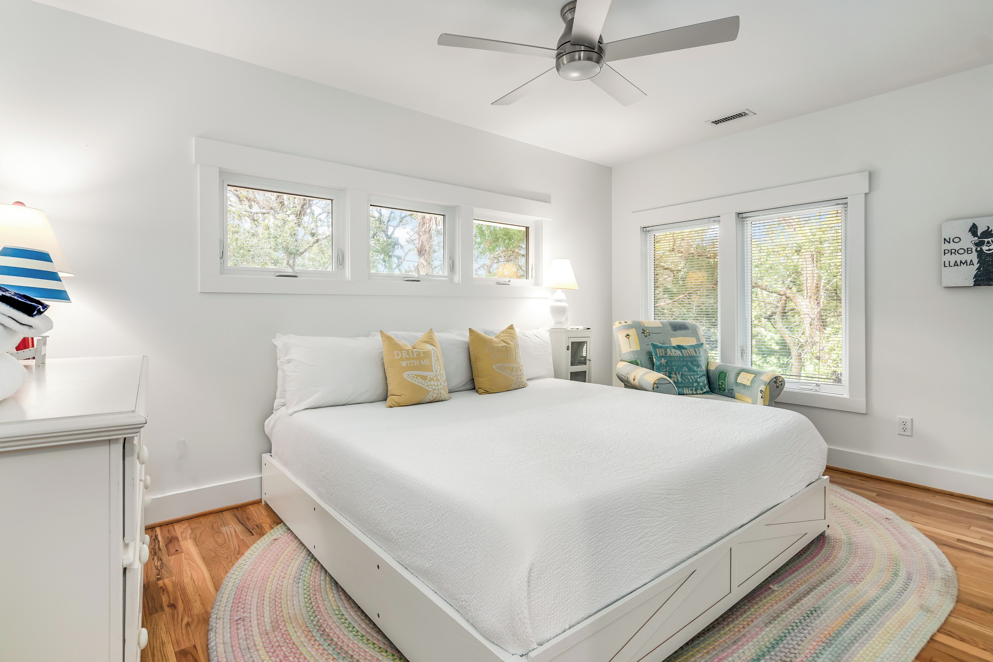 A bedroom with a white bed, multiple windows, a ceiling fan, and a bedside lamp.