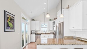 A modern kitchen with white cabinets, a granite countertop, and a large island with bar stools. - Aqua Vista | Oceanfront 5BR w/ Private Pool, Hot Tub & Stunning Views (Emerald Isle)