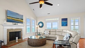A spacious living room with a fireplace, a large flat-screen TV, a leather sofa, and a glass coffee table. - Aqua Vista | Oceanfront 5BR w/ Private Pool, Hot Tub & Stunning Views (Emerald Isle)