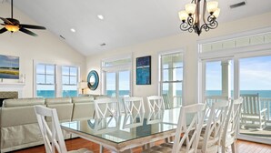 A spacious dining area with a glass table and white chairs, large windows with ocean views, a ceiling fan, and a chandelier. - Aqua Vista | Oceanfront 5BR w/ Private Pool, Hot Tub & Stunning Views (Emerald Isle)