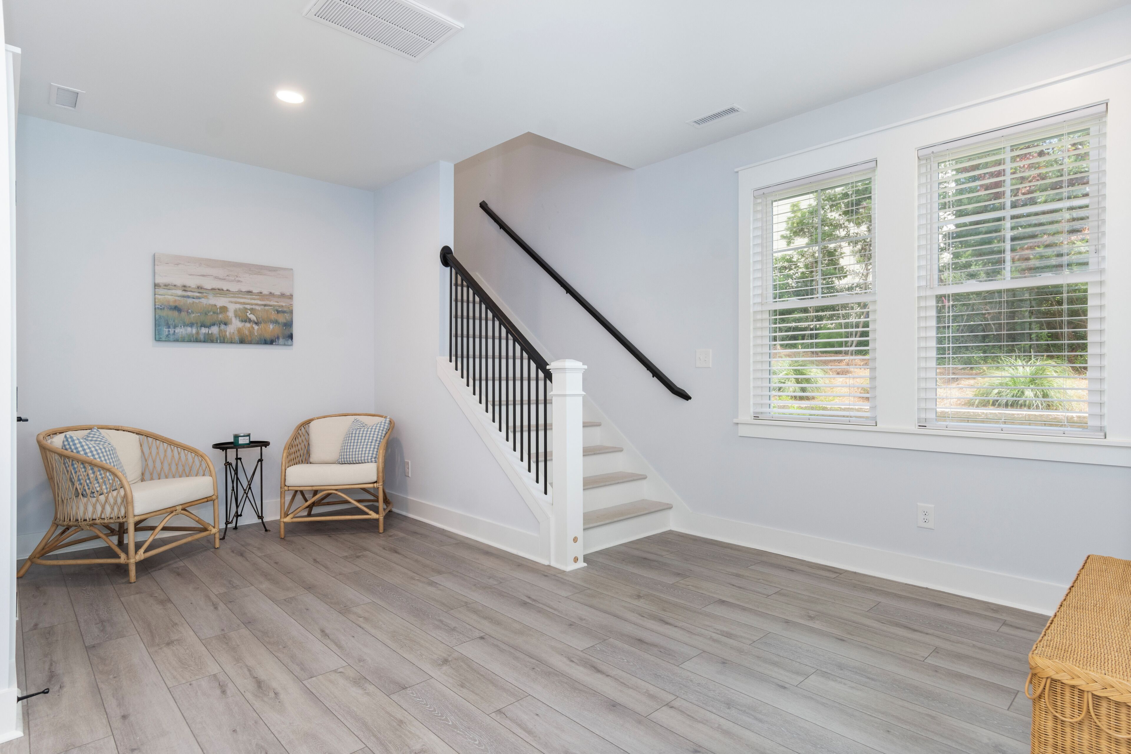 A spacious room with a wooden floor, a white staircase, two wicker chairs, a small table, and a large window with blinds.