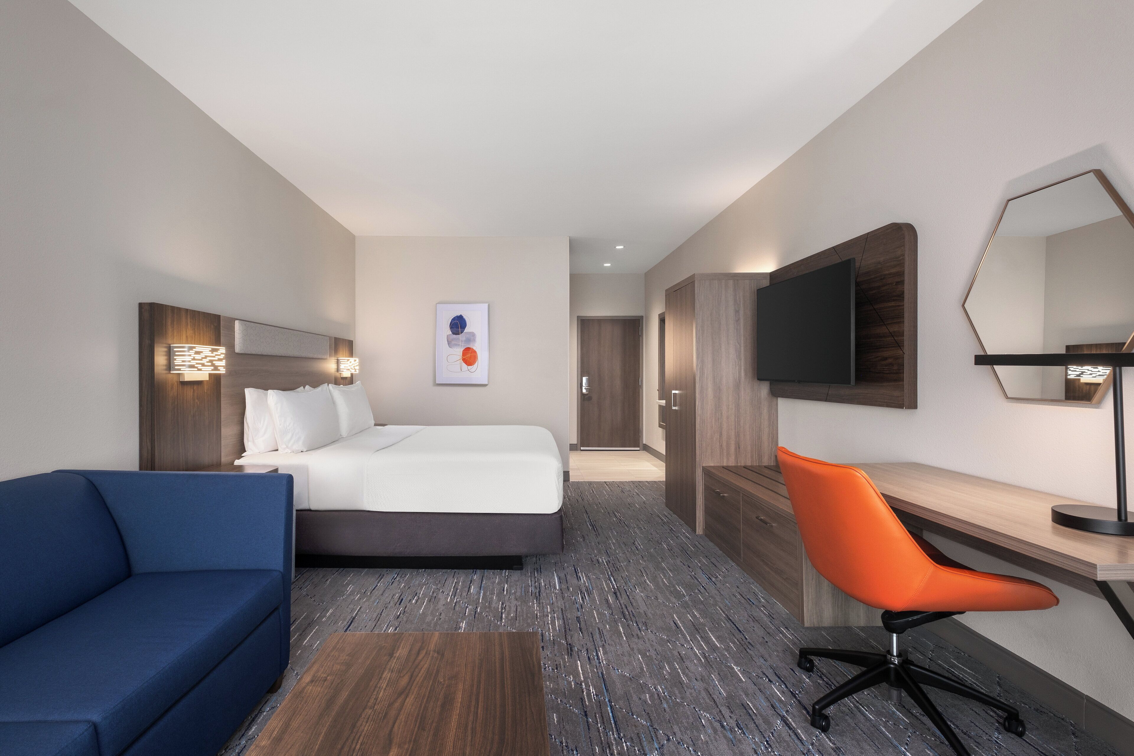 Ein modernes Hotelzimmer mit einem Bett, einem blauen Sofa, einem orangefarbenen Bürostuhl, einem Holztisch, einem großen Spiegel und einem an der Wand befestigten Fernseher.
