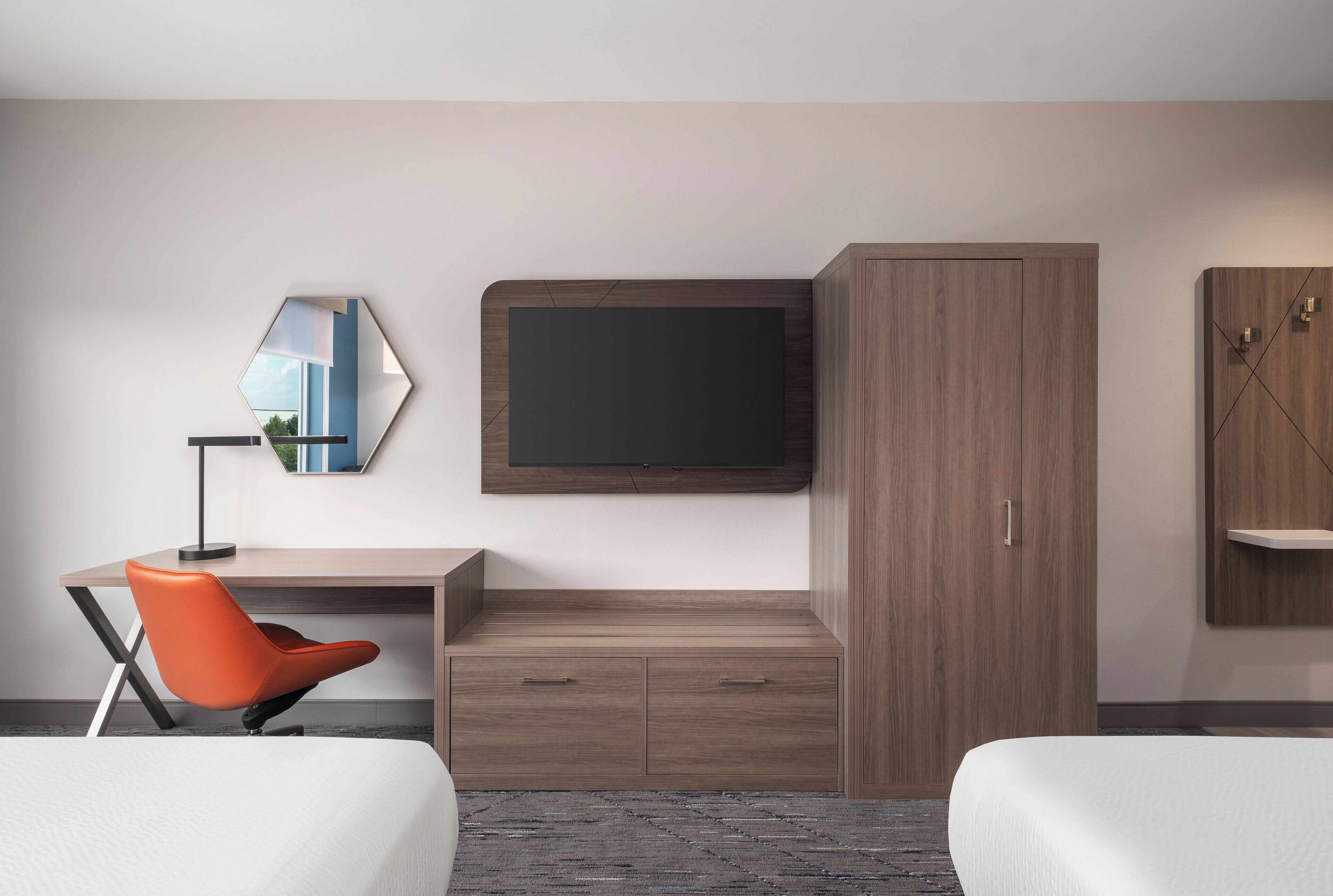 A hotel room with a bed, a desk with a chair, a television mounted on the wall, and a wardrobe.