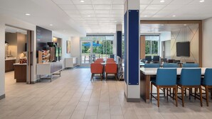 A modern dining area with a long counter and blue chairs, a kitchen area with a refrigerator and coffee station, and large windows offering a view of the outdoors. - Holiday Inn Express & Suites Buford – Mall of Georgia by IHG (Buford)