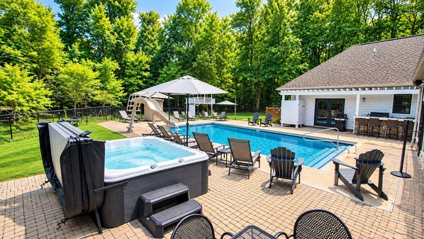 A backyard with a swimming pool, hot tub, lounge chairs, and a covered seating area.