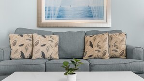 Ein graues Sofa mit gemusterten Kissen, ein weißer Couchtisch und ein gerahmtes Bild von einer Meereslandschaft.