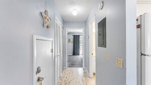 Ein Flur mit hellblauen Wänden, einer weißen Tür mit Schlüsselloch, einer Deckenleuchte und einem grauen Teppich, der zu einem Raum mit Fenster und Vorhang führt.