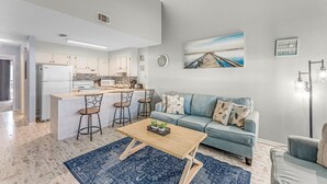 Eine moderne Küche mit einer zentralen Kochinsel, ein Essbereich mit einem Holztisch und ein Wohnzimmer mit Sofa und Gemälde.