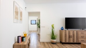 Una sala moderna con piso de madera, un televisor sobre un mueble de madera, una planta y un banco con recipientes de colores.