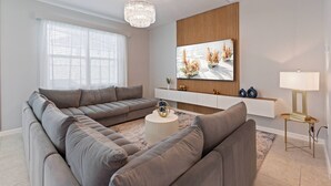 Een moderne woonkamer met een hoekbank, een ronde salontafel en een flatscreen televisie bevestigd aan een houten wandpaneel.