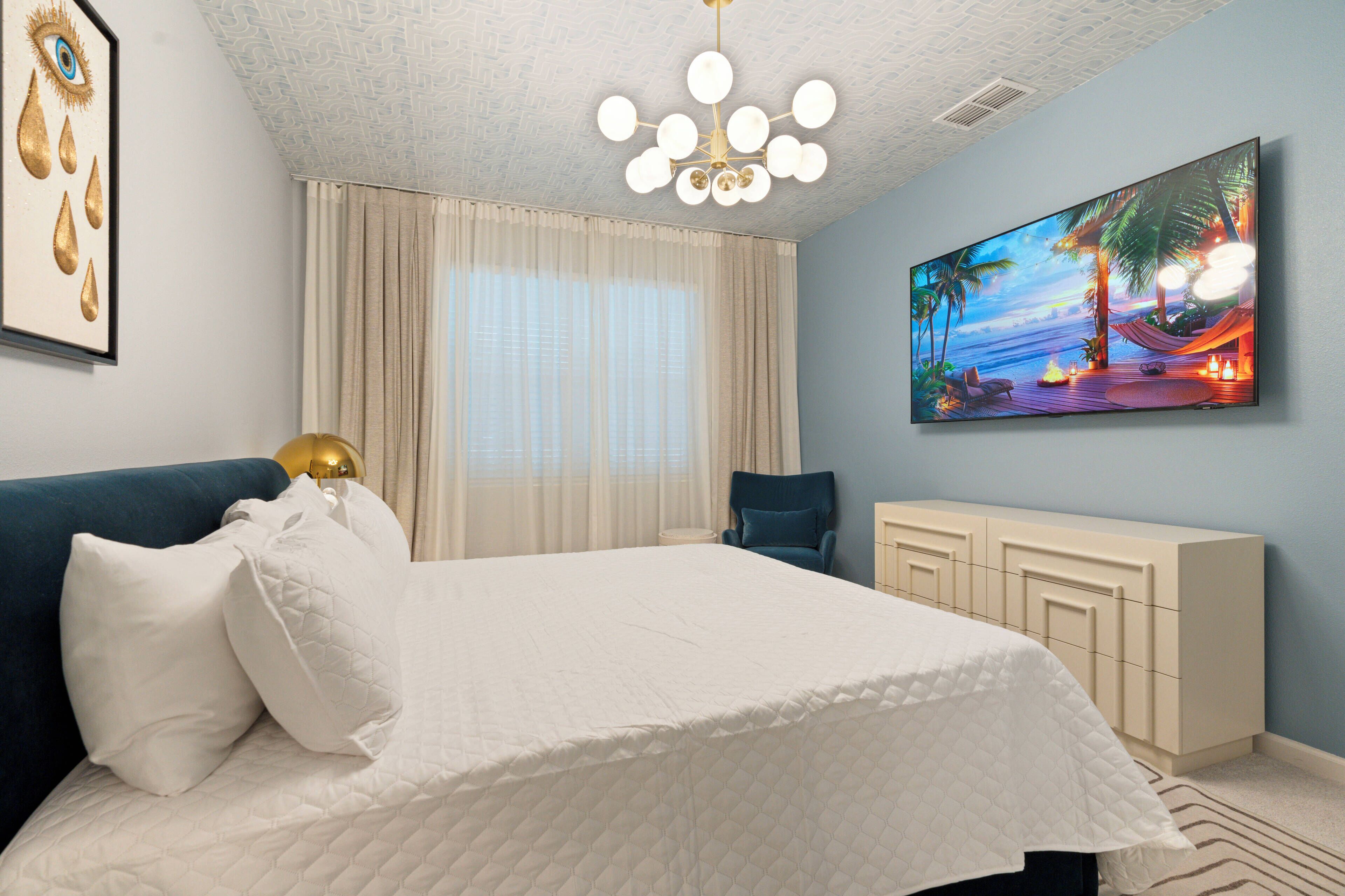 A bedroom with a bed, a nightstand, a chair, a TV, and a painting of a beach scene.