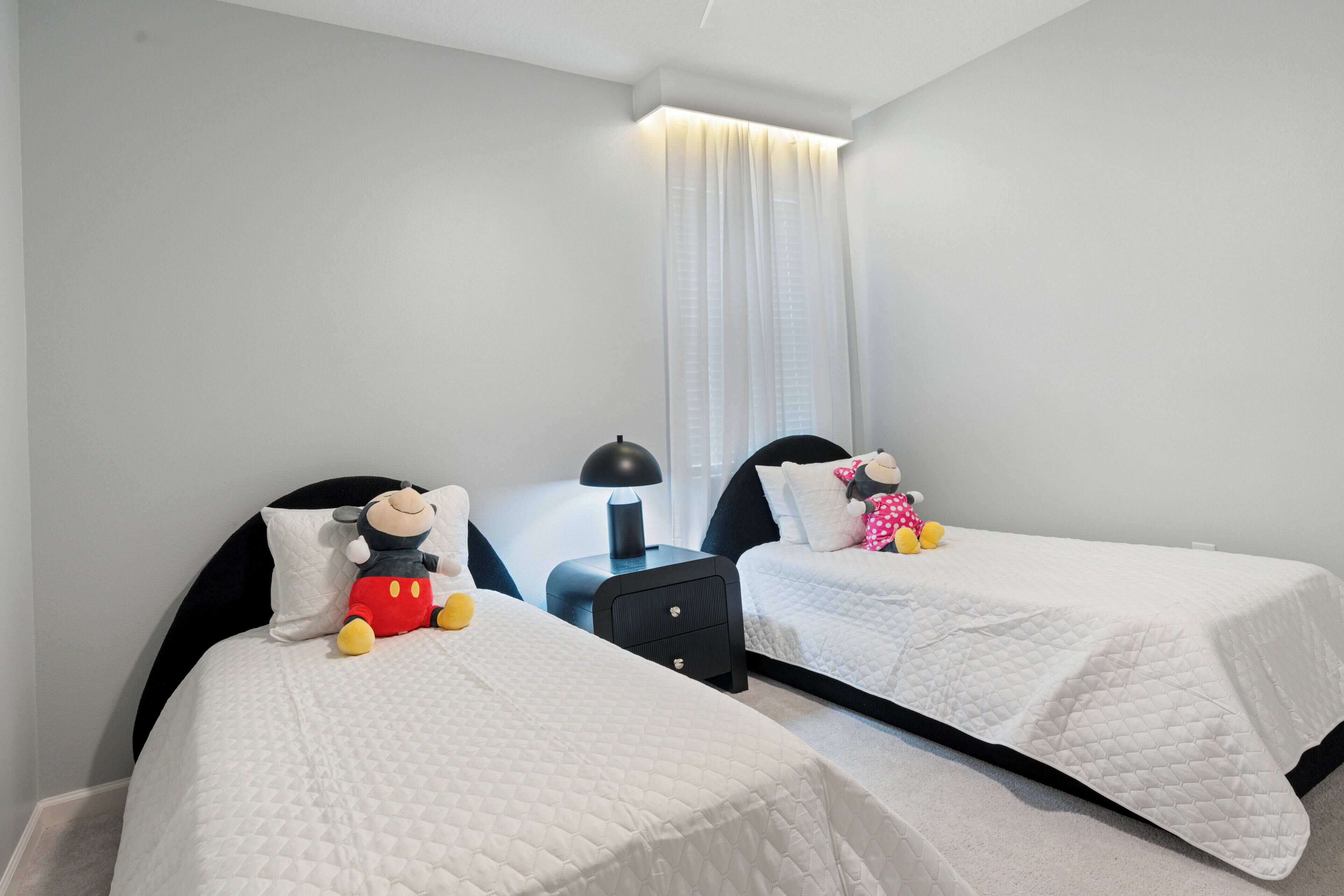 Un dormitorio con dos camas, cada una con una colcha blanca acolchada y un peluche de Mickey Mouse. Entre las camas hay una mesita de noche negra con una lámpara.