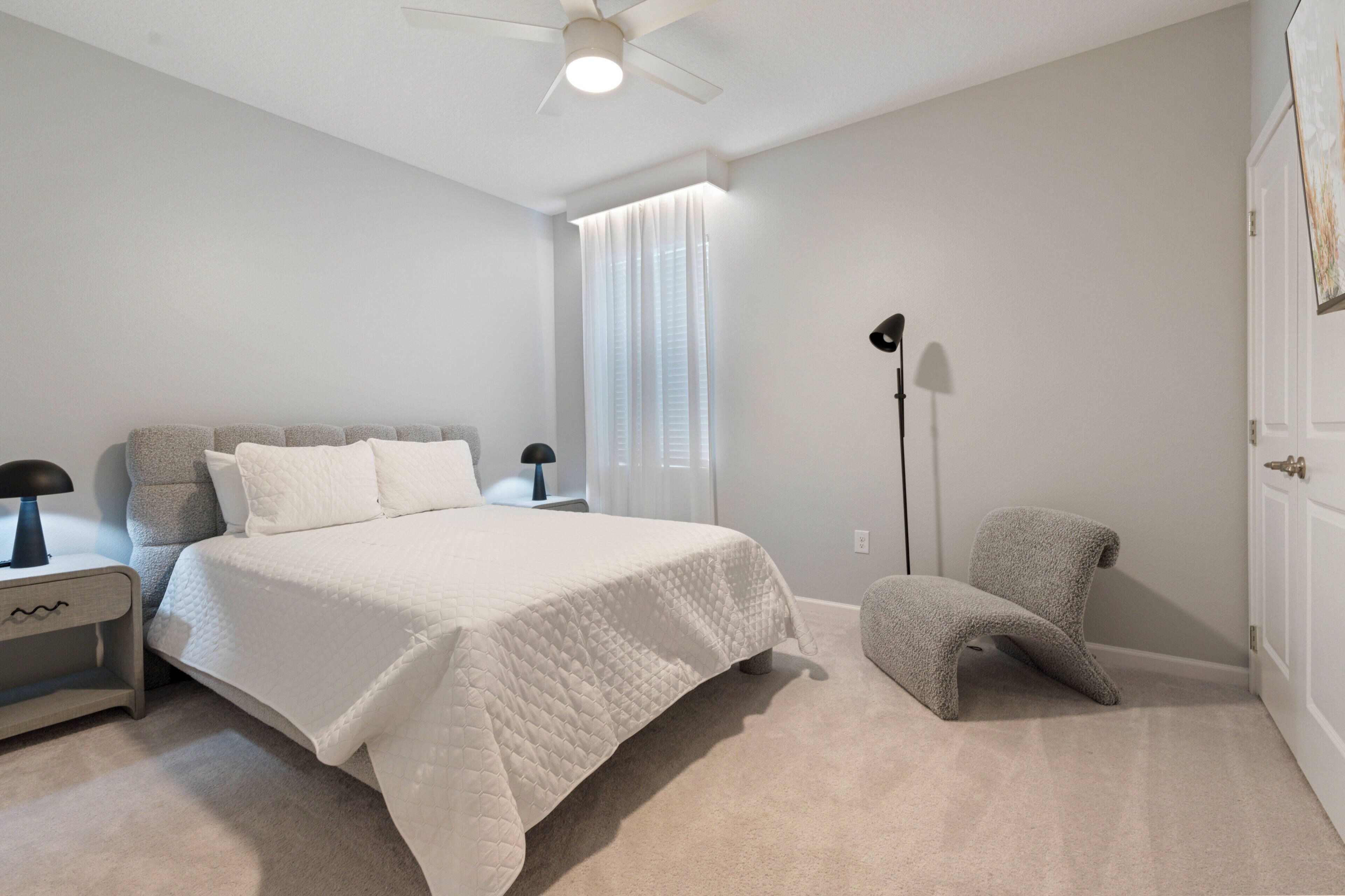 Unwind in this cozy bedroom with a plush bed dressed in crisp white linens and soft gray tones. Sheer curtains allow natural light to filter in, creating a tranquil atmosphere ideal for relaxation and peace.