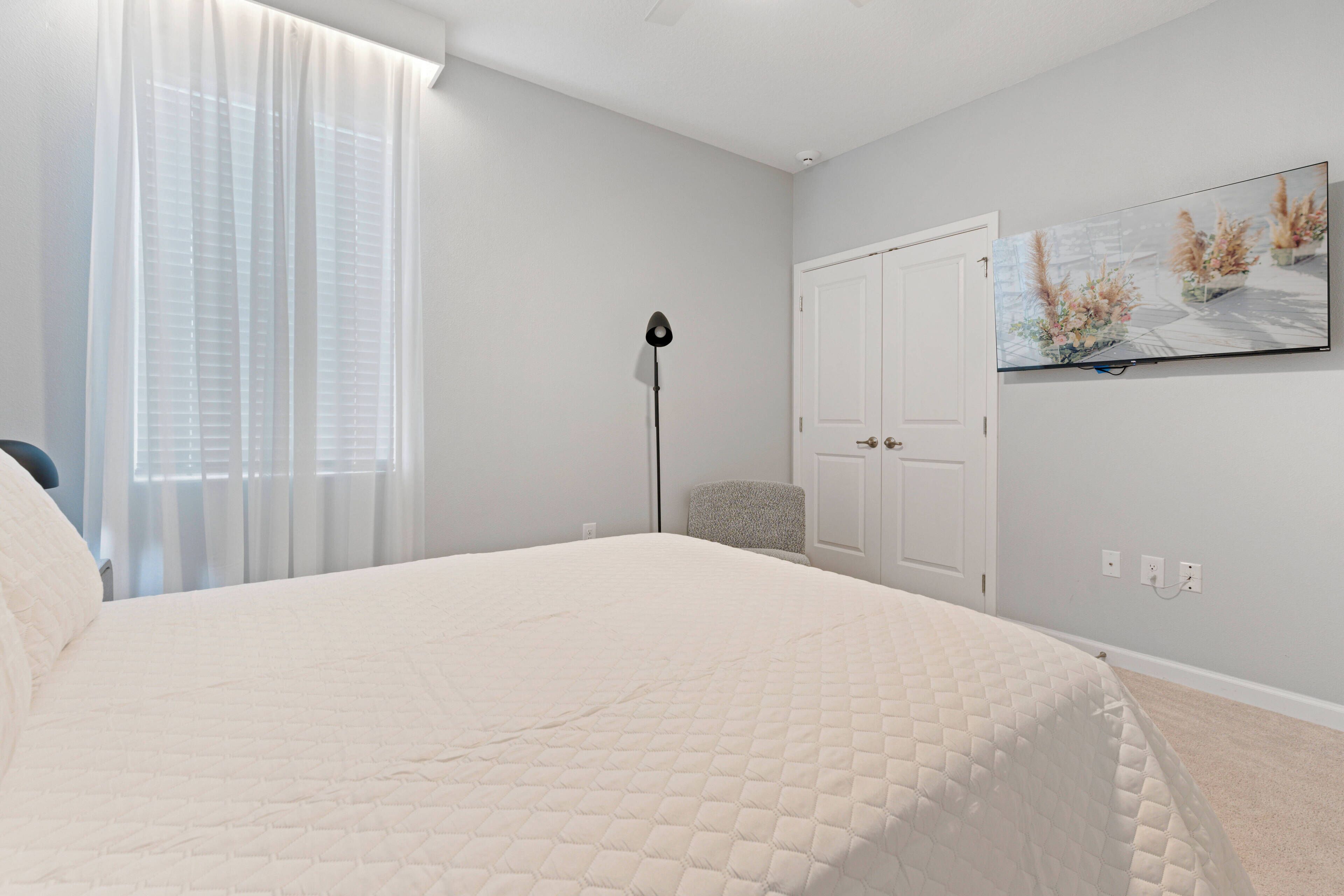 A bedroom with a bed, a chair, a floor lamp, a door, and a wall-mounted TV.