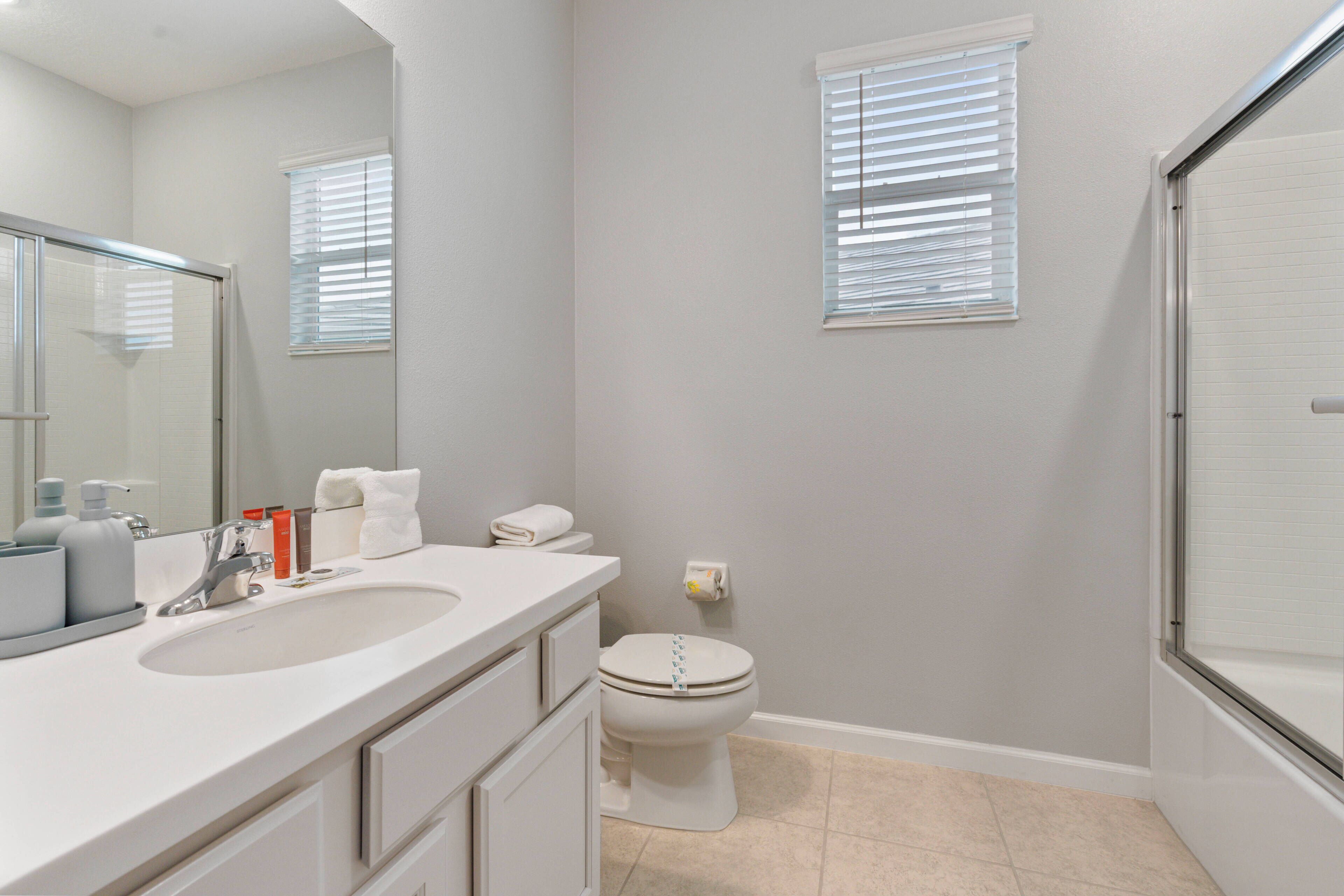 A bathroom with a white sink, mirror, and toilet. There are two windows with blinds, and a shower area is visible.