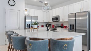 A modern kitchen with white cabinets, a stainless steel refrigerator, a large island with bar stools, and a hanging light fixture.