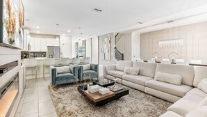 Una sala de estar moderna con sofá, mesa de centro y televisor. En el fondo, se observa una cocina con armarios y un refrigerador.