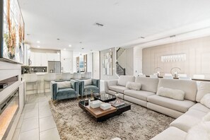 Uma sala de estar moderna com sofá, mesa de centro e televisão. Cozinha visível ao fundo, com armários e geladeira.
