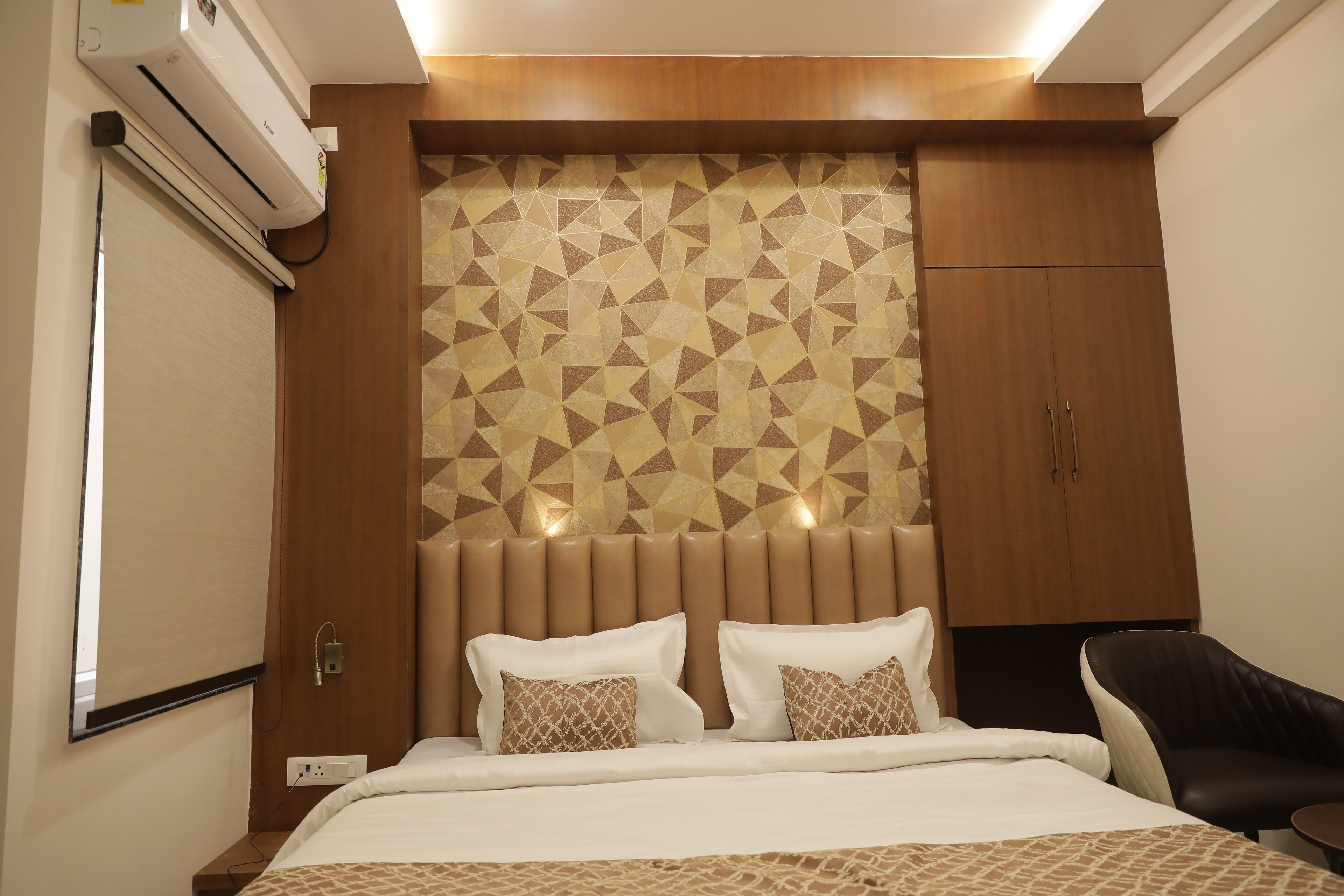 Habitación de hotel con cama, cabecero de madera, pared con estampado geométrico, sillón de cuero negro y armario de madera.
