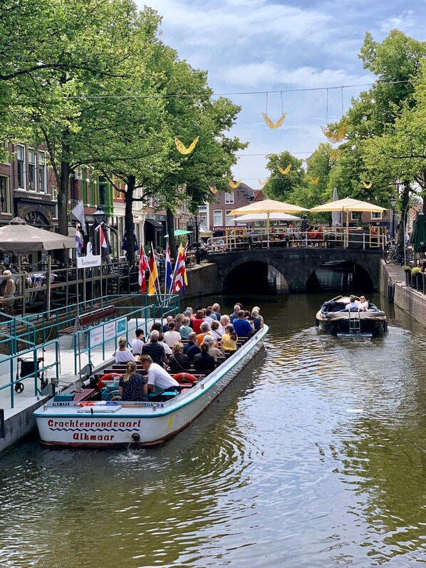 A boat with passengers on a canal, a bridge, and buildings with flags.