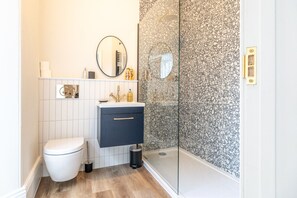 Une salle de bain moderne avec des toilettes, un lavabo et un meuble sombre, un miroir rond et une douche en verre avec un mur recouvert de carreaux à motifs.