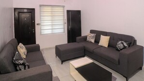 A modern living room with a sectional sofa, a coffee table, and a large window with blinds. - Charming 2-Bedroom House in Ajah with AC & WiFi (ajah)