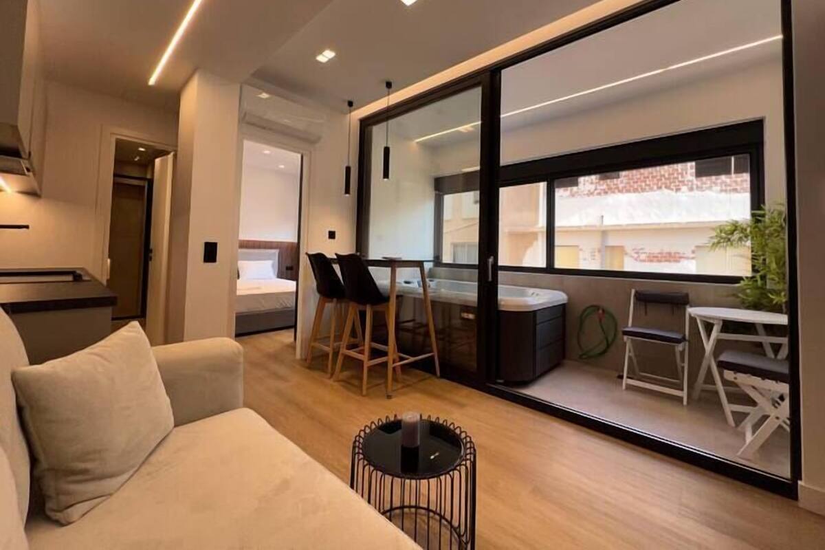 A modern hotel room with a large sliding glass door leading to a balcony, a sofa, a small round table, and a dining area with chairs.