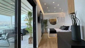A modern hotel room with a large window, a bed, a desk, and a view of the city. - BlueMotion - CentralSuite (Piraeus)