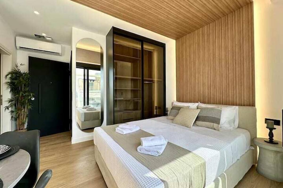 A modern bedroom with a large bed, a desk, and a chair. There is a plant, a wardrobe, and a window.