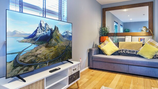 A modern living room with a large flat-screen TV on a white stand, a grey sofa with yellow and grey cushions, a wooden floor, and a large window with blinds. - Queens Quarter 2-Bed | Next to City Centre - Pass the Keys (Belfast)