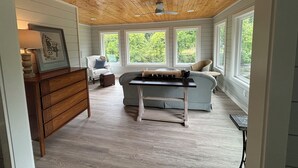 A room with a wooden ceiling, large windows, a sofa, a table, and a dresser.